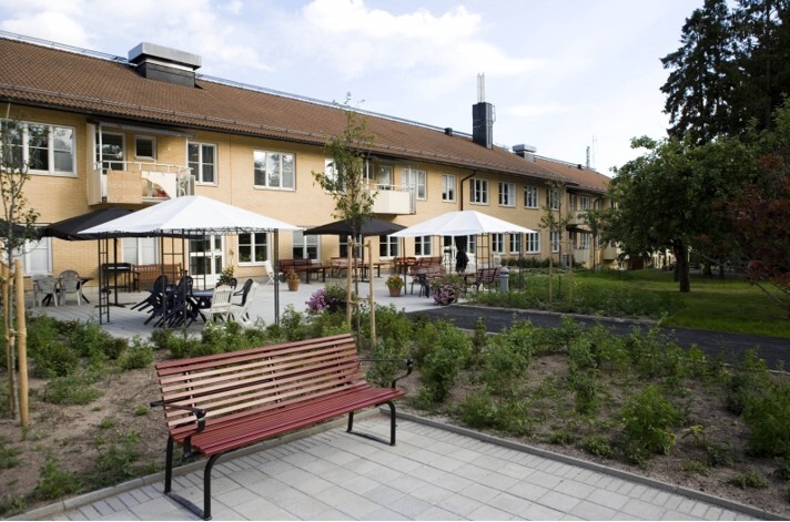 En park med en låg gul byggnad i bakgrunden; Brommagården där Ebab bytte ventilationsaggregat och byggde miljöhus.